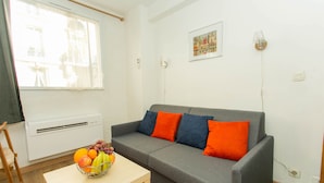 Een woonkamer met een grijze bank, oranje en blauwe kussens, een houten tafel met een fruitschaal, een witte radiator en een raam met dunne gordijnen.