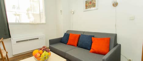 Un salon avec un canapé gris, des coussins orange et bleus, une table en bois sur laquelle se trouve un panier de fruits, un radiateur blanc et une fenêtre ornée de rideaux légers.