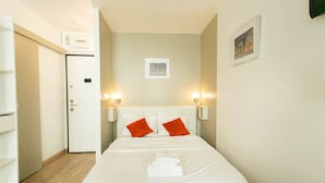 Een net opgemaakt bed met witte beddengoed en twee oranje kussens, een witte kledingkast, en een deur met lichtschakelaar.
