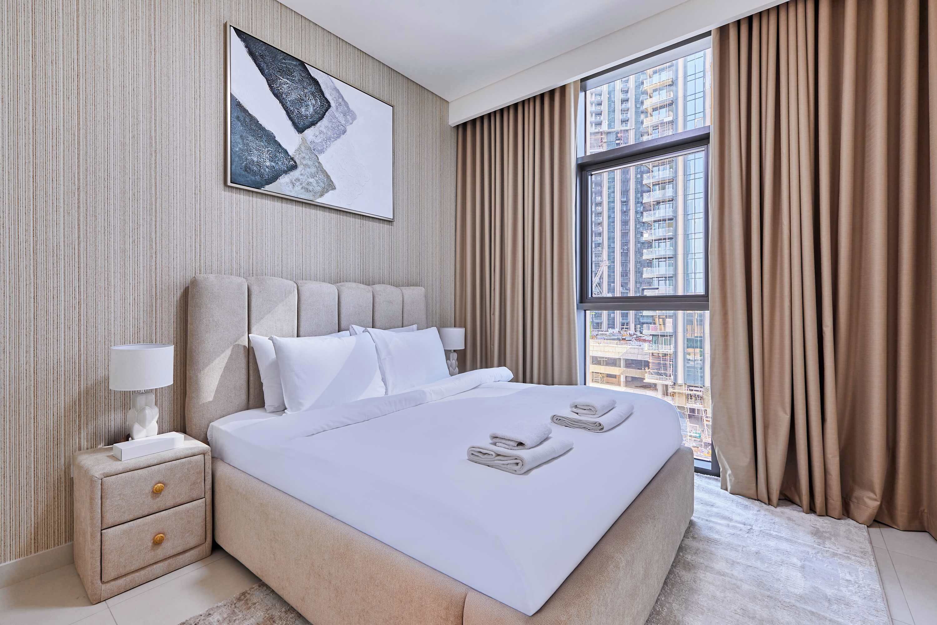 A modern hotel room with a large bed, a nightstand with a lamp, a framed artwork on the wall, and a view of the city through a large window.