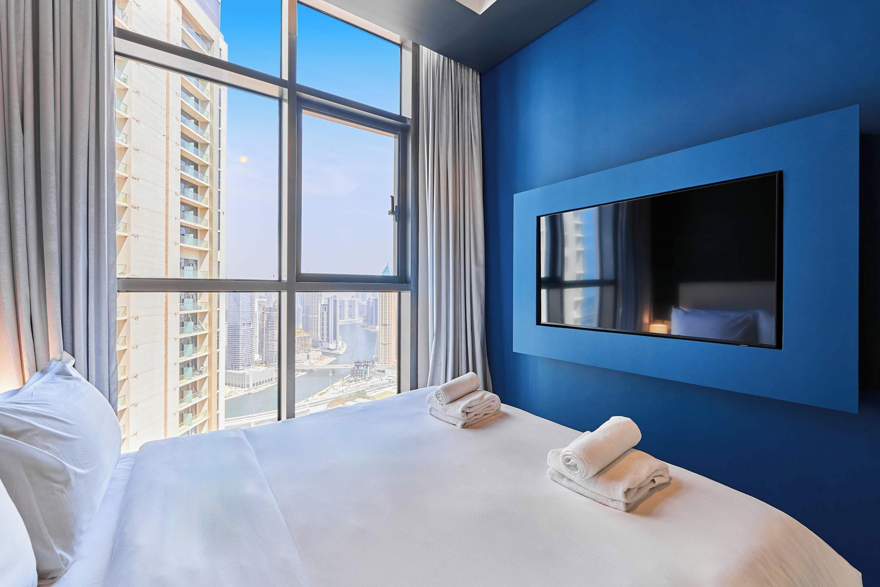 A hotel room with a large bed, a flat-screen TV mounted on the wall, and a view of a cityscape through a large window.