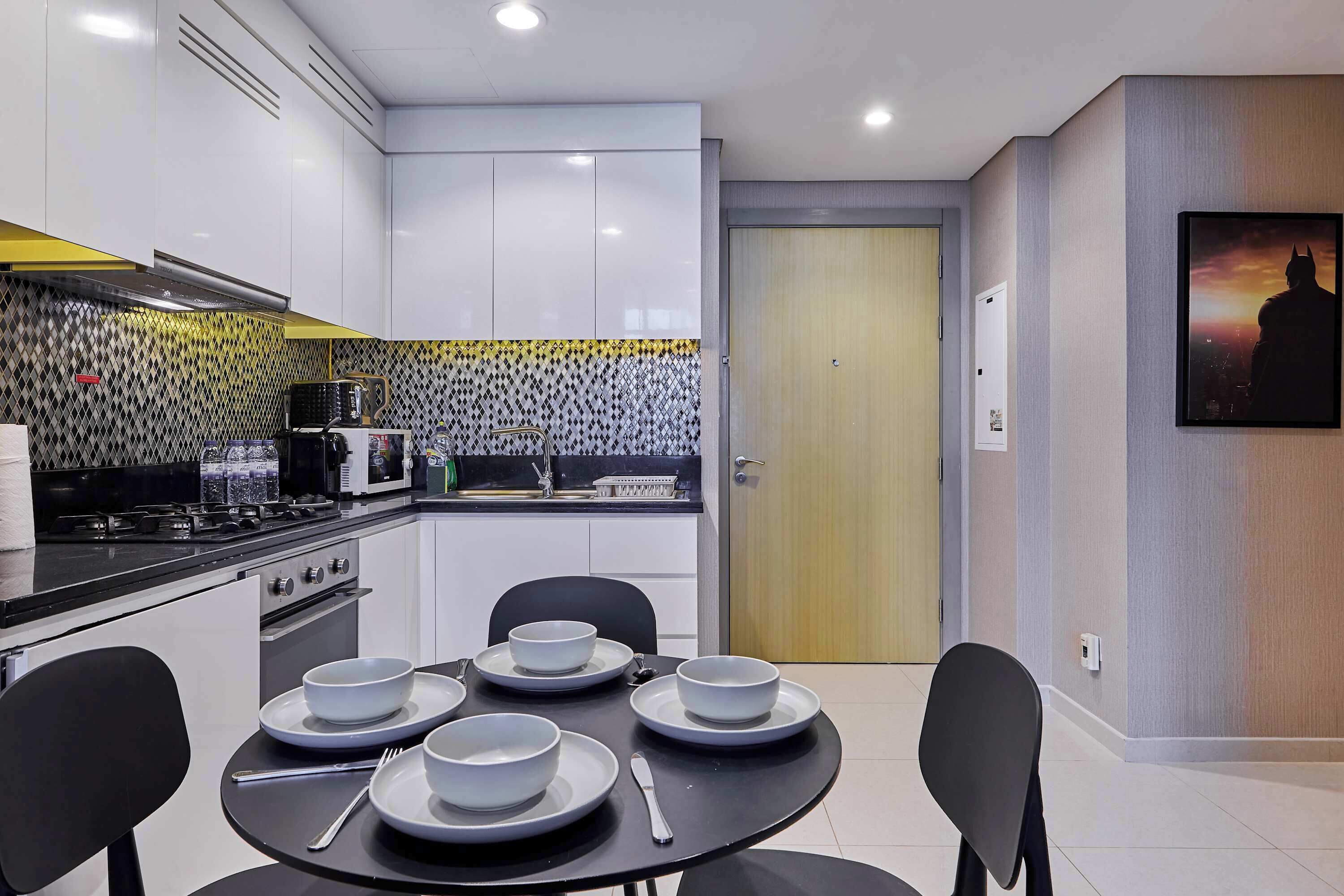 A modern kitchen with white cabinets, a black countertop, and a patterned backsplash. There is a dining table with plates and cutlery.