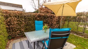 Een terras buiten met een glazen tafel, blauwe stoelen en een gele parasol.