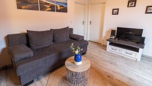 A living room with a grey sofa, a round coffee table with a vase, a flat-screen TV on a white cabinet, and a wall with three framed pictures. - Klara vacation apartment in Greetsiel - quiet and in the middle of it all (Greetsiel)
