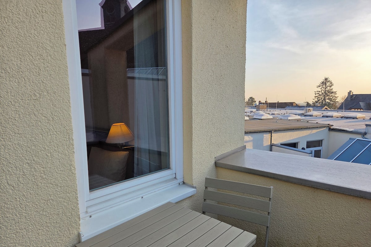 Un balcón con una mesa y una silla, una ventana con una lámpara y vistas a tejados.