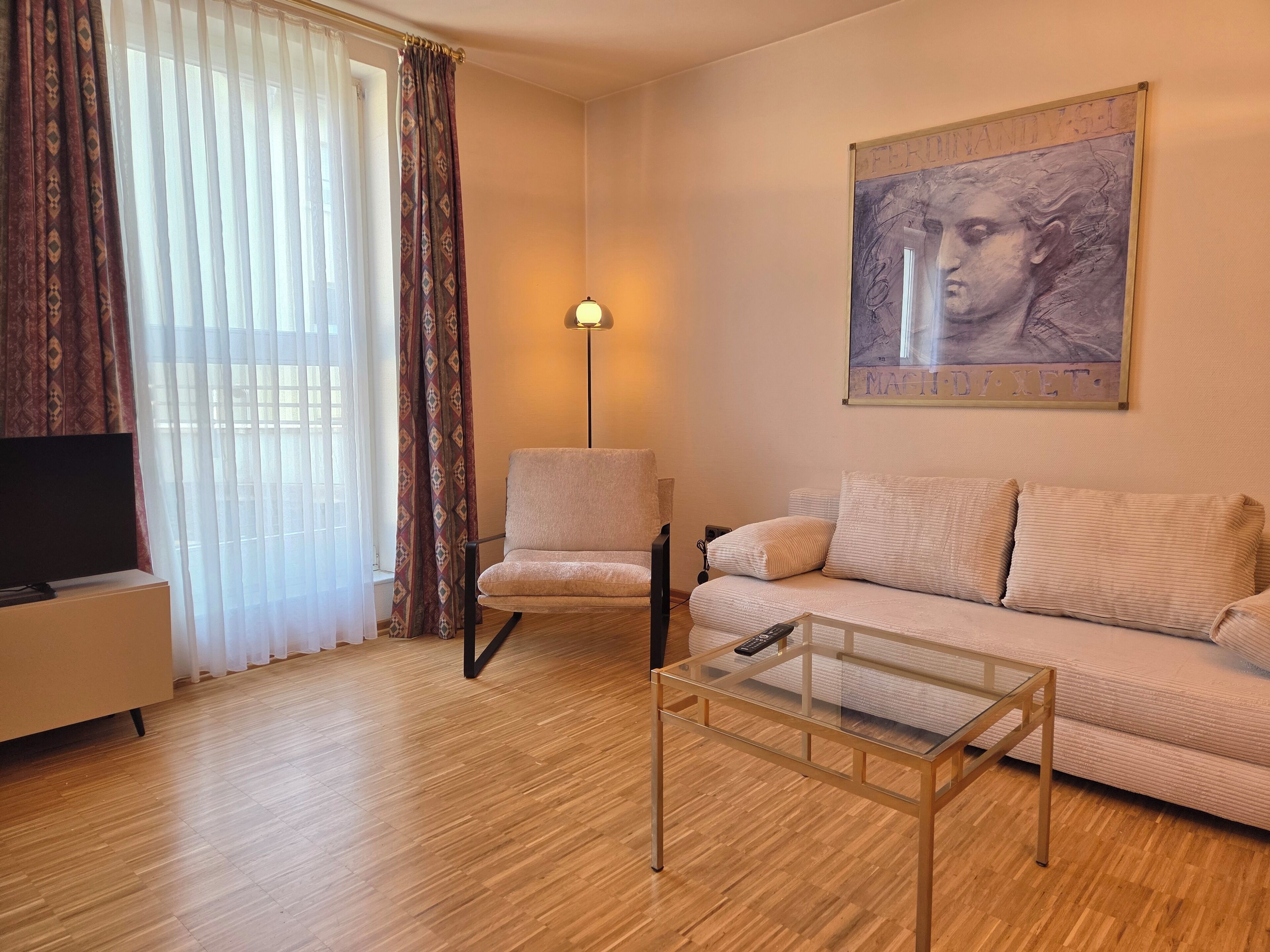 Ein Wohnzimmer mit einer Couch, einem Glastisch, einem Sessel, einem Fernseher und einem Bild an der Wand.