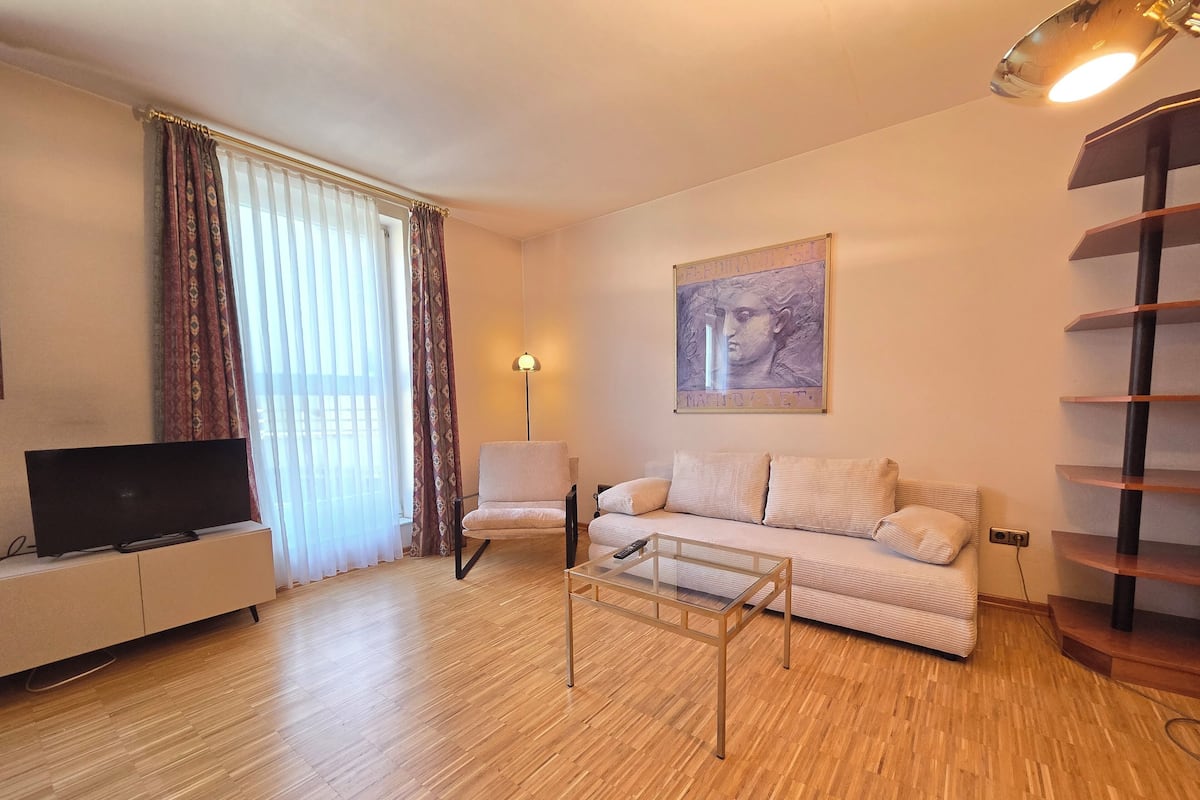 Una sala de estar con un sofá, una silla, un televisor sobre un mueble bajo, una lámpara y un cuadro en la pared.