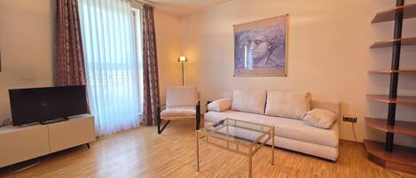 Una sala de estar con un sofá, una silla, un televisor sobre un mueble bajo, una lámpara y un cuadro en la pared.