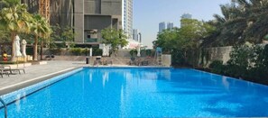 Ein Swimmingpool mit Liegestühlen und Sonnenschirmen, umgeben von Grünflächen und hohen Gebäuden.