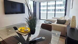 A modern living room with a glass dining table, a sofa, and a TV mounted on the wall.
