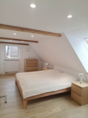 Une chambre avec un plafond en pente, un lit recouvert d’une couverture blanche à texture, une commode en bois, une table de chevet avec une lampe et une fenêtre donnant sur un immeuble.