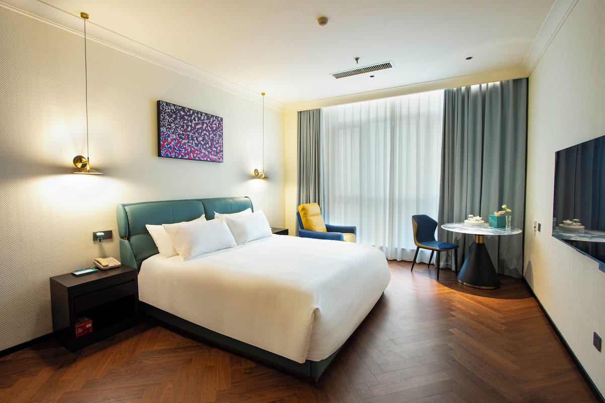 A hotel room with a large bed, bedside table with a phone, a desk with a chair, and a large window with curtains.