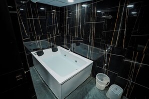 A modern bathroom with a large rectangular bathtub, black marble walls with gold veins, and a small trash bin. - Hotel The Avanta - Gandhinagar (Gandhinagar)