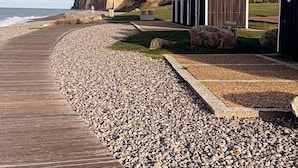 A coastal walkway with a wooden boardwalk, pebbles, and a row of beach huts. - Le Pré Marin (Sotteville-sur-Mer)