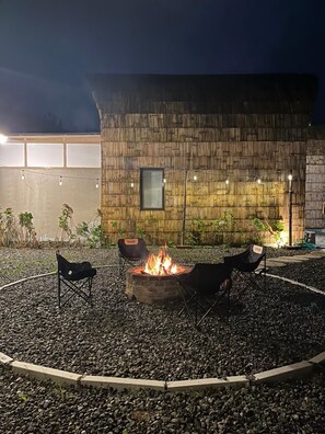 Un foyer avec deux chaises, entouré de galets, adossé à un mur en bois et illuminé de guirlandes lumineuses.