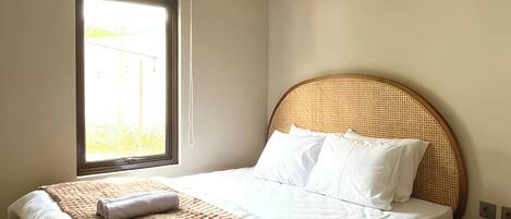 Ein ordentlich bezogenes Bett mit Holz-Kopfteil, ein Fenster mit Jalousien und ein Nachttisch mit gewebtem Überzug.