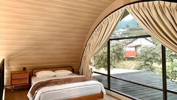 Un dormitorio con una cama grande, una mesita de noche de madera, un ventilador de techo y vistas al exterior a través de una ventana grande con cortinas.