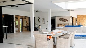 Ein geräumiger Wohnbereich mit Esstisch für vier Personen, umgeben von Korbstühlen. Deckenventilatoren, ein großes Fenster und Blick auf einen Pool sind vorhanden.