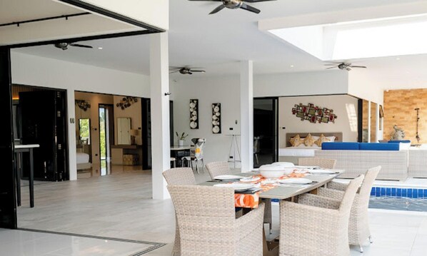 Ampla sala de estar com mesa de jantar para quatro pessoas, rodeada por cadeiras de vime. Há ventiladores de teto, uma janela grande e vista para uma piscina.