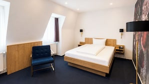 A hotel room with a bed, a blue chair, two bedside tables, a nightstand, a window with curtains, and a painting on the wall. - DORMERO Hotel Crailsheim (Crailsheim)