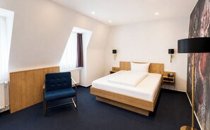 A hotel room with a bed, a blue chair, two bedside tables, a nightstand, a window with curtains, and a painting on the wall. - DORMERO Hotel Crailsheim (Crailsheim)