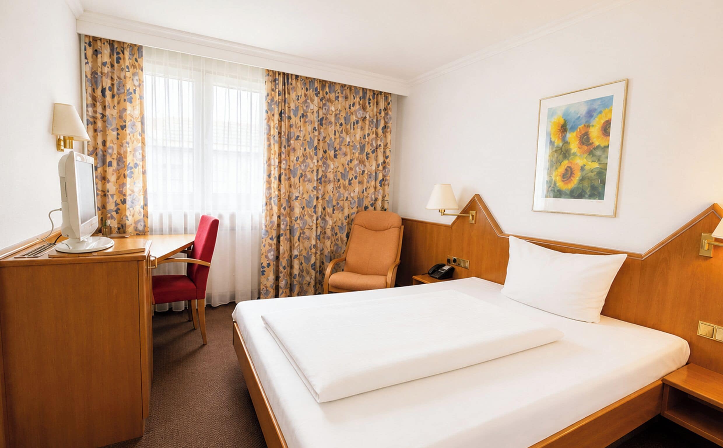 A hotel room with a bed, desk, chair, and a painting on the wall.