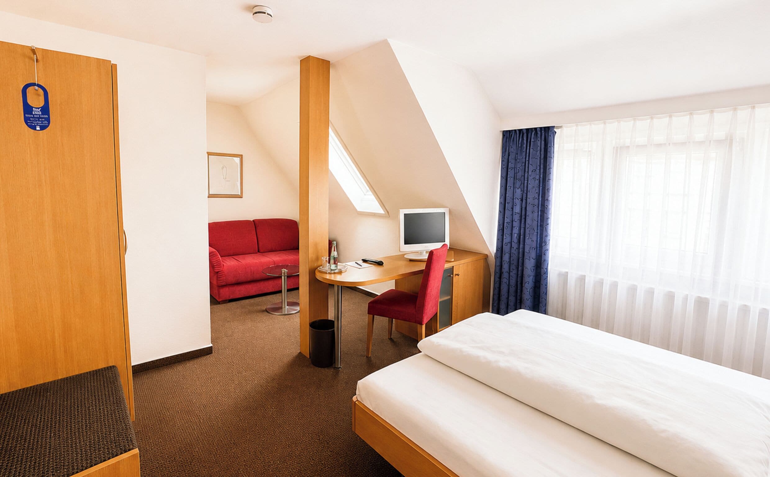 Habitación de hotel con cama, escritorio con computadora, sofá rojo y ventana con cortinas.