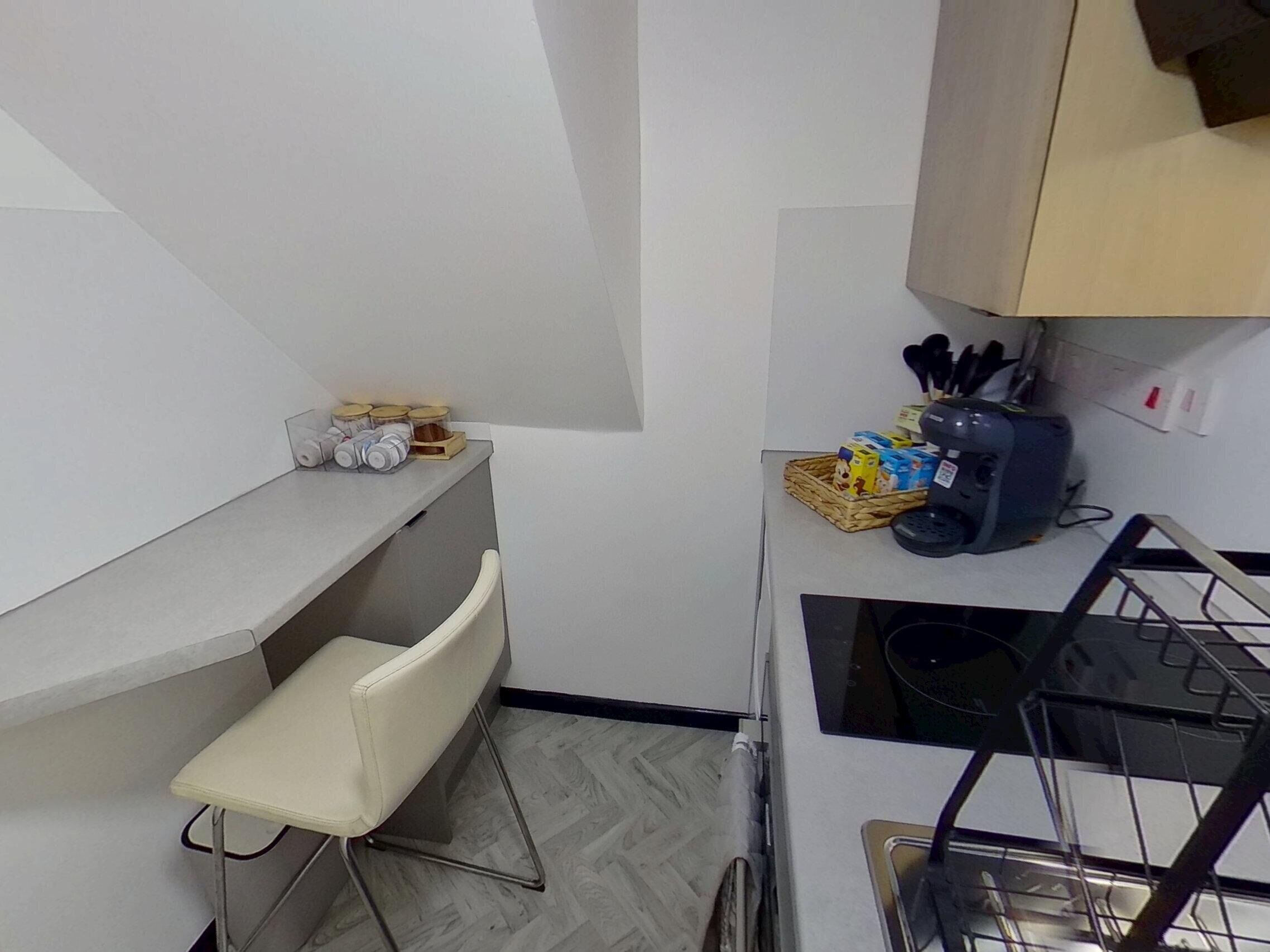 A compact kitchen with a white countertop, a small chair, a sink, and a stove. There are jars and a basket on the countertop.