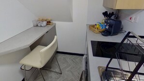 Pieni, käytännöllinen keittiö, jossa on valkoinen työtaso, pieni tuoli, pesuallas ja liesi. Työtasolla on purkkeja ja kori.