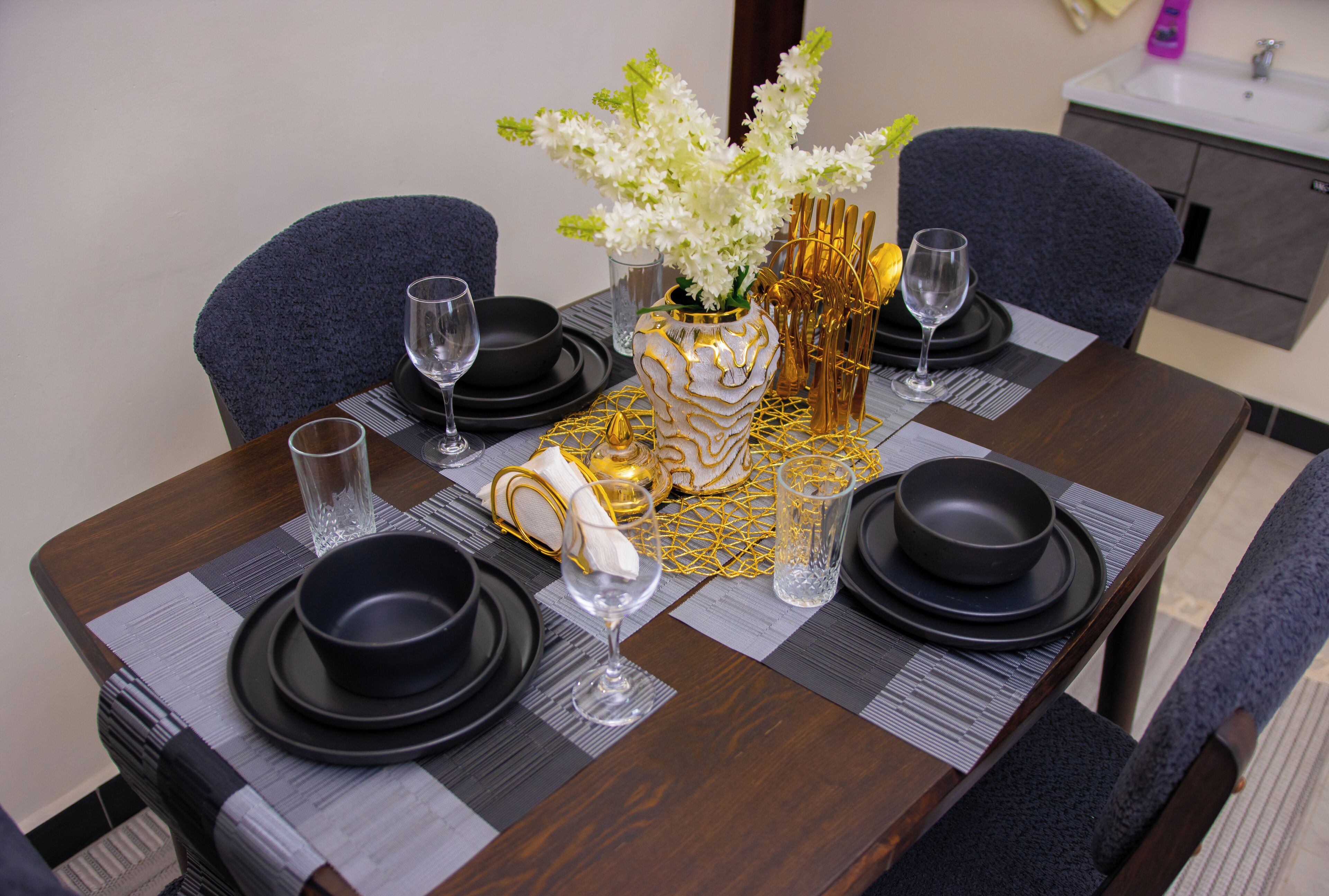 Une table à manger dressée avec des assiettes noires, des verres transparents et un vase contenant des fleurs blanches.