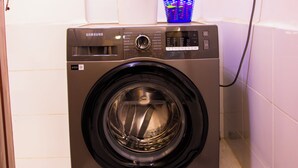 Une machine à laver Samsung à chargement frontal, installée dans une salle de bain aux murs et au sol recouverts de carrelage blanc.