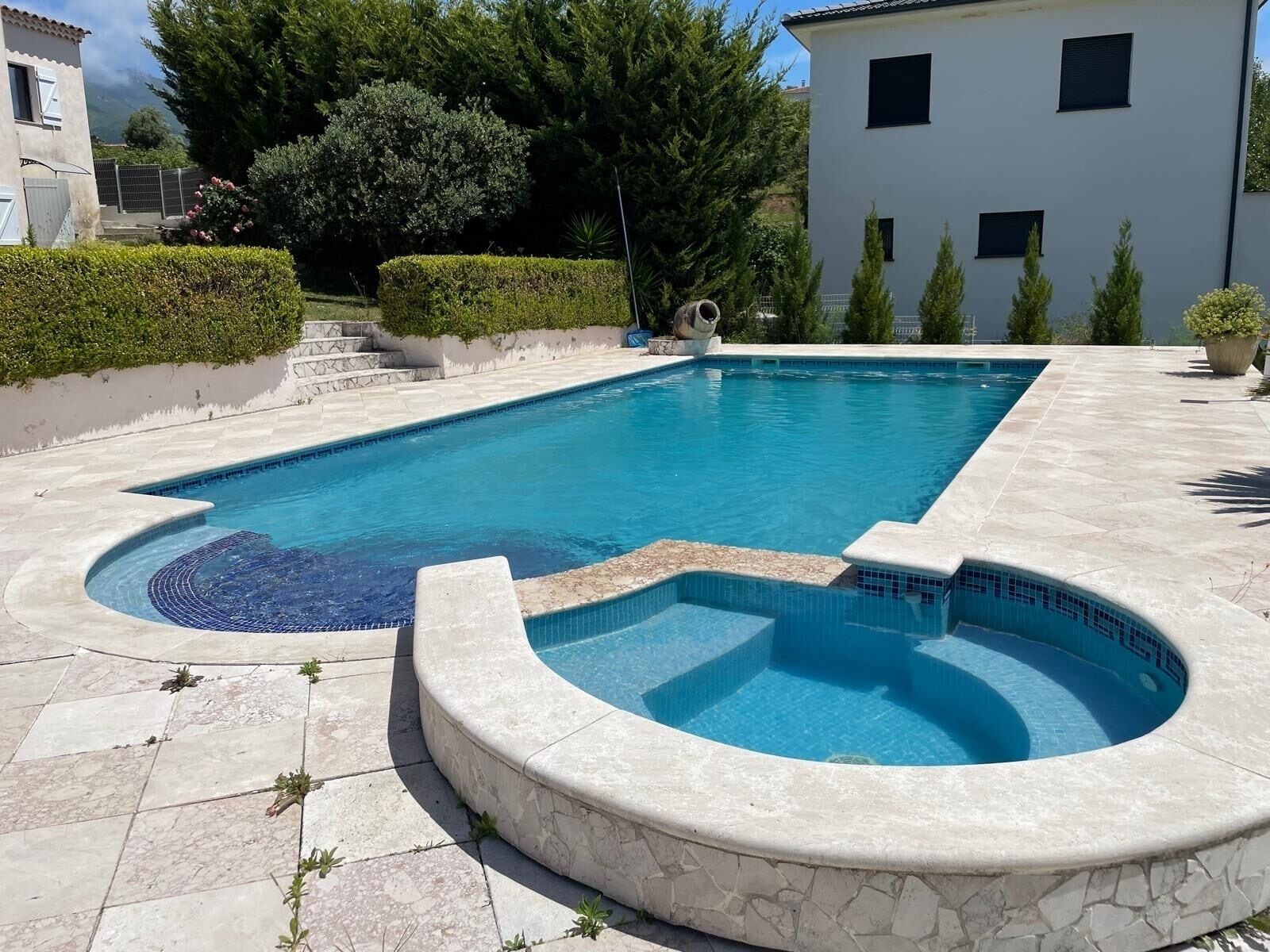 Ein rechteckiger Pool mit einer kleinen Whirlwanne, umgeben von Steinplatten und Grünpflanzen.