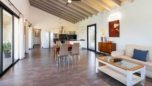 A spacious living area with a dining table, chairs, a sofa, and a coffee table. A kitchen is visible in the background. - 2 Cortons - Wall (Muro)