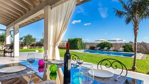 An outdoor dining area with a view of a swimming pool, palm trees, and a well-maintained lawn. - 2 Cortons - Wall (Muro)