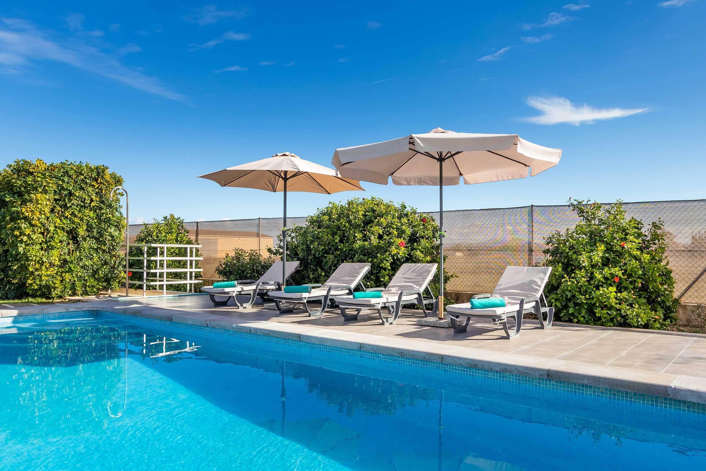 A swimming pool with lounge chairs and umbrellas, surrounded by greenery and a fence.