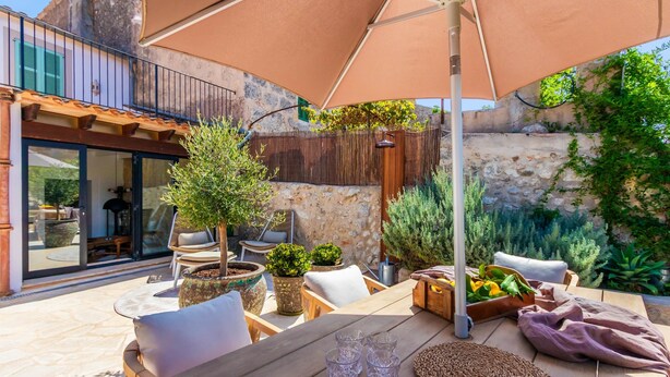 An outdoor patio with a wooden table, chairs, and a large umbrella. There are potted plants and a stone wall in the background. - Can Miguel (Sineu)