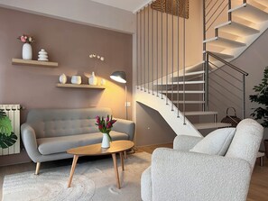 A modern living room with a grey sofa, a round wooden coffee table, a white armchair, a staircase with a metal railing, and a wall-mounted shelf with decorative items. - Chianti's Secret Nest - Cozy Home with Sunset View (Impruneta)