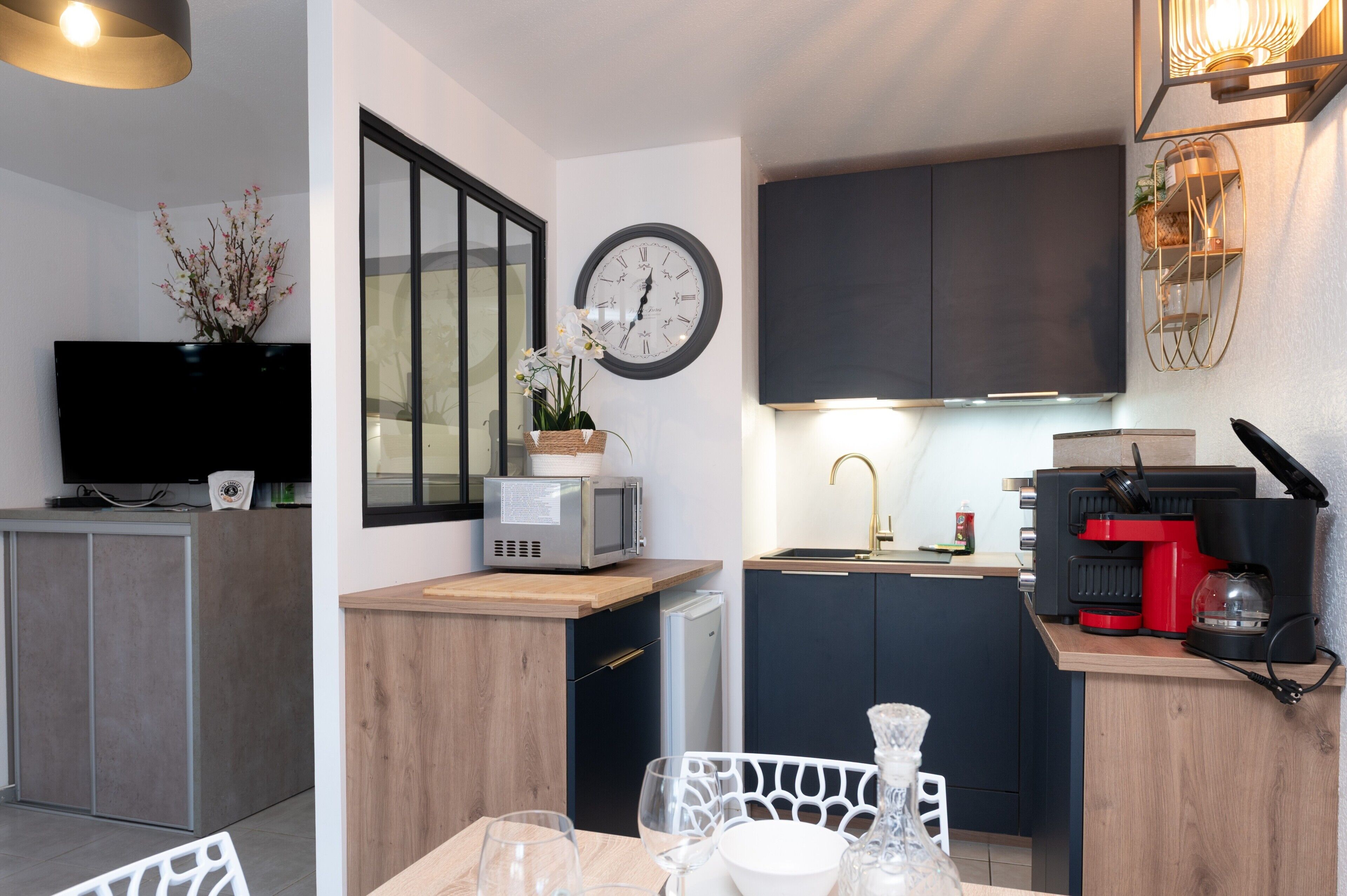 Eine kompakte Küche mit Mikrowelle, Toaster und Kaffeemaschine, ein Essbereich mit Tisch für zwei Personen und eine an der Wand montierte Uhr.