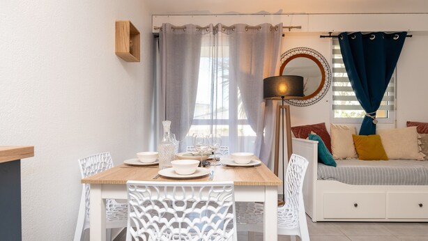 A dining area with a wooden table and white chairs, a bed with a blue curtain, and a wall-mounted lamp. - Ty Avel Apartment with Shared Pool, Balcony, and Wi-Fi (Quiberon)