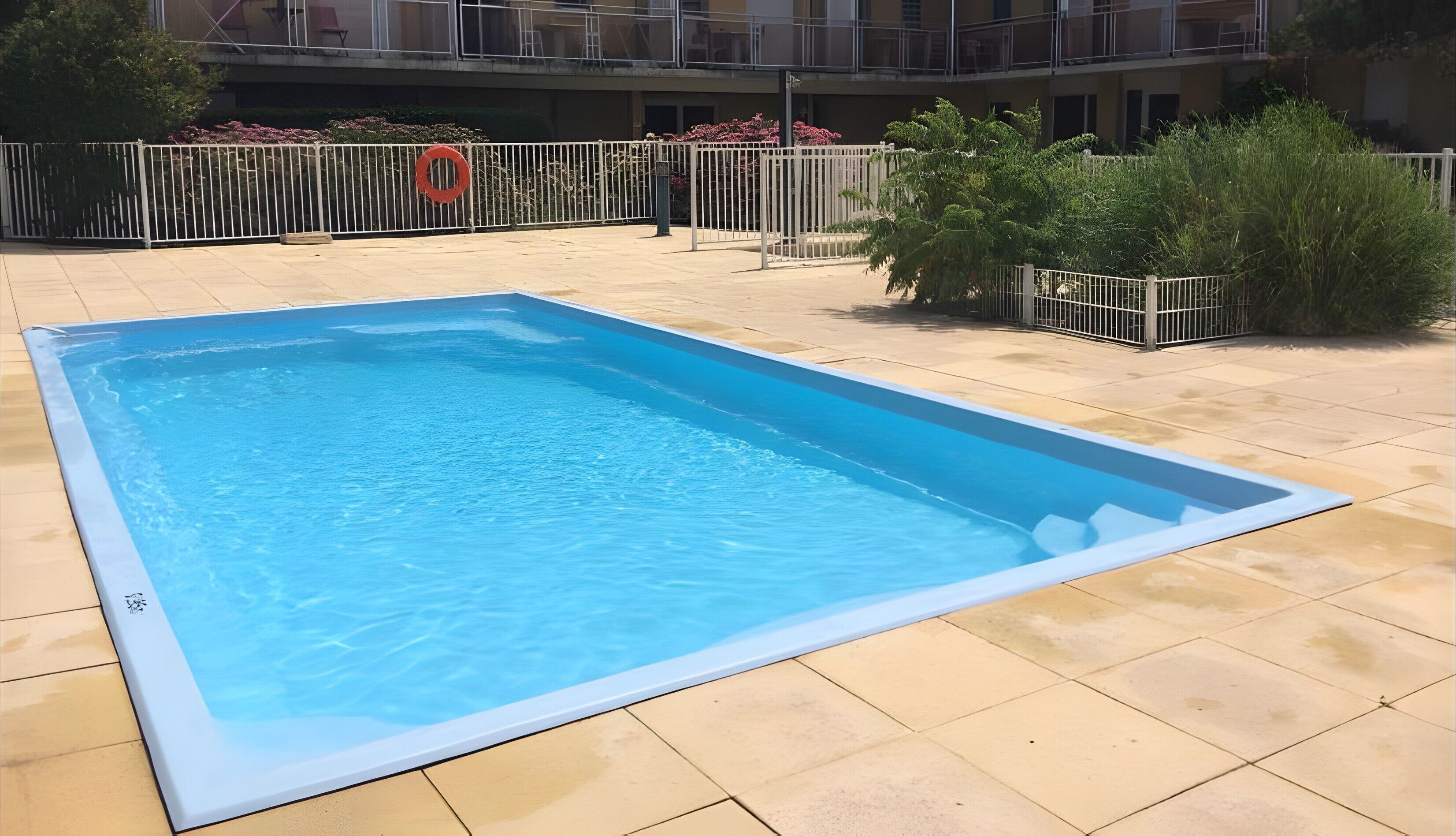 Una piscina rectangular con borde de baldosas, rodeada por una cerca metálica y un flotador salvavidas.