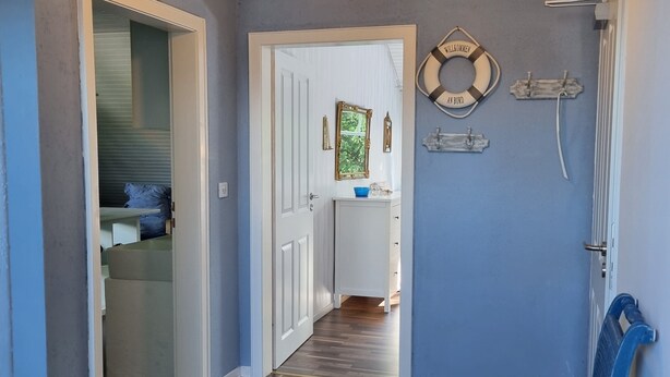 Een gang met een houten vloer, een witte deur, een klein tafeltje met een blauwe kom, en een aan de muur bevestigd reddingsboei met tekst.