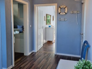 Ein Flur mit Holzboden, einer weißen Tür, einem kleinen Tisch mit einer blauen Schale und einem an der Wand befestigten Rettungsring mit Text.