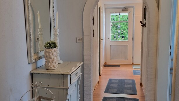 Ein Flur mit einer gewölbten Tür, die in einen Raum mit Fenster führt. Links befindet sich ein Spiegel, ein Schminktisch mit einer Blumenvase und eine Tür mit Griff.