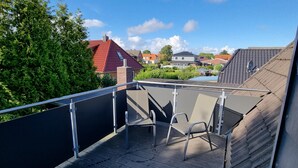 Una terrazza sul tetto con tavolo e sedie, circondata da una ringhiera metallica, con vista su un quartiere residenziale con case e alberi.