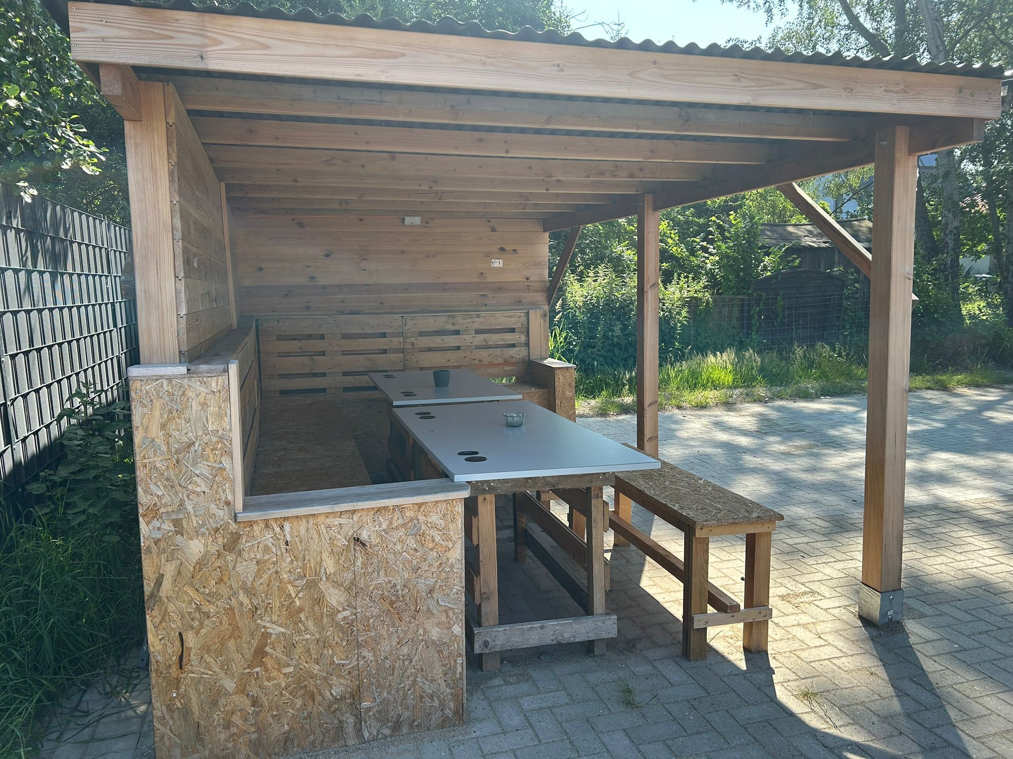 A wooden outdoor kitchen structure with a table and benches, surrounded by a metal fence and greenery.