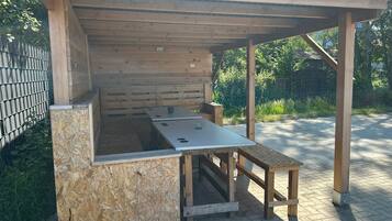 A wooden outdoor kitchen structure with a table and benches, surrounded by a metal fence and greenery.