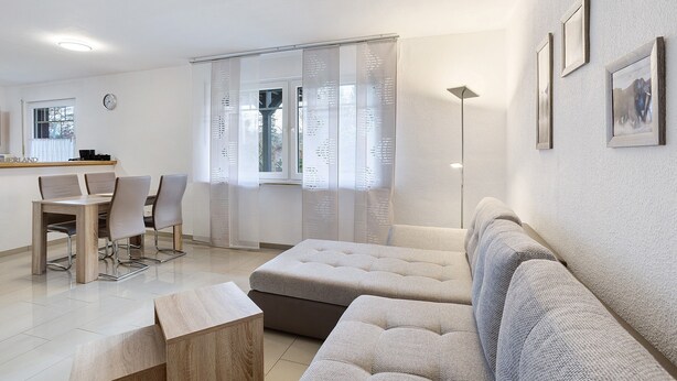 Ein modernes Wohnzimmer mit einem Ecksofa, einem Esstisch mit Stühlen und einem großen Fenster mit leichtem Vorhang.
