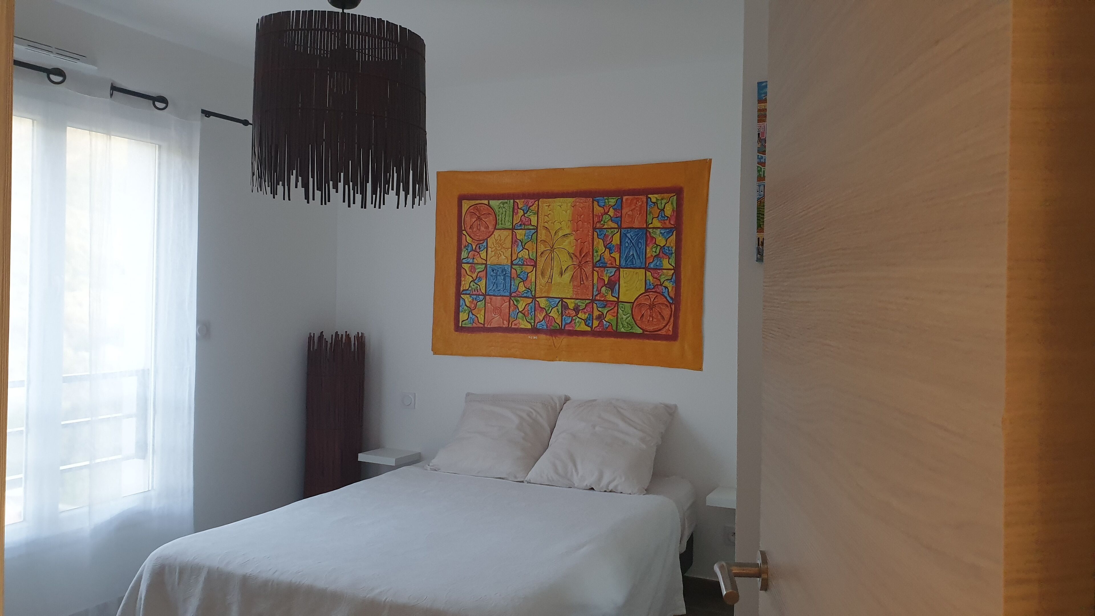 A bedroom with a bed, a hanging light fixture, a colorful tapestry, and a window with curtains.