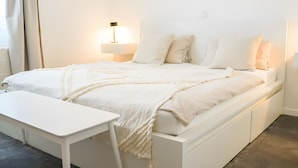 A white bed with a bedside table, a lamp, and a wall-mounted light fixture. - Holiday Apartment Im Alten Feld (Schmallenberg)