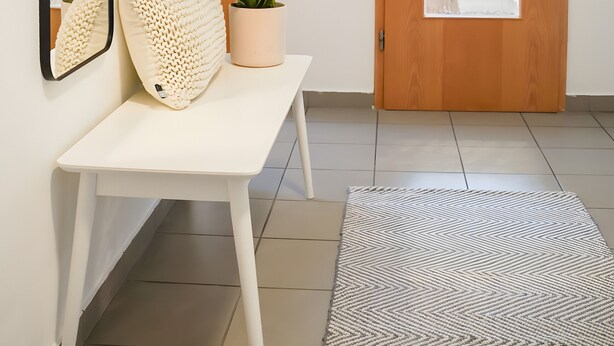 A hallway with a wooden door, a white bench with cushions, a potted plant, a mirror, and a patterned rug. - Holiday Apartment Im Alten Feld (Schmallenberg)